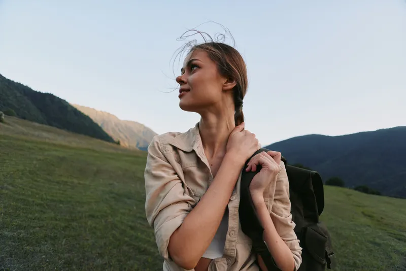Young woman with a backpack walks across a grassy hillside, looking ahead with a calm, hopeful expression under a clear blue sky during a peaceful outdoor excursion.