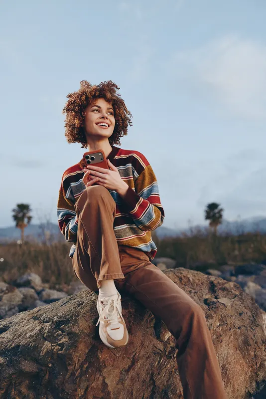 Woman lifestyle outdoors portrait with curly hair, striped sweater, sitting on rock, holding smartphone, relaxed mood, mountains and blue sky.