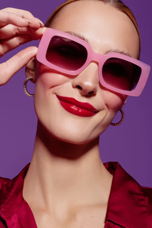 Close-up of confident woman wearing large pink sunglasses and red lipstick against purple background. Fashion concept with bold accessories and vibrant colors.