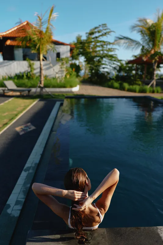 A woman rests at the edge of a tropical pool, hands behind her head, eyes closed, soaking in sun. Palm trees frame a bright resort scene with sparkling water and a calm moment of leisure.