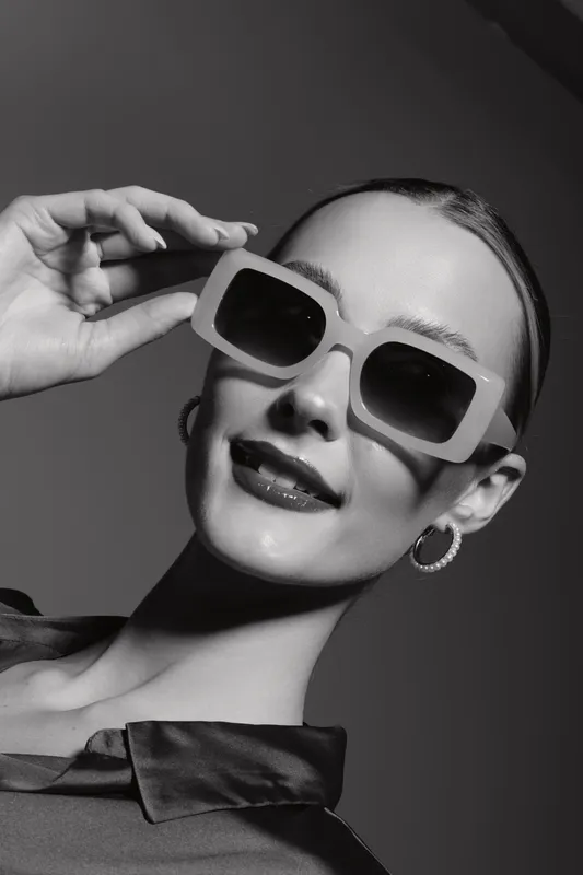 Young white woman with pale skin wearing large sunglasses smiles confidently in a studio portrait with dark background.