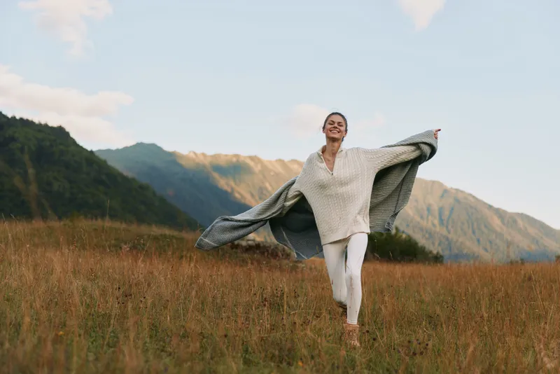 Happy woman in cozy sweater running through grassy field with mountains in background. Concept of freedom, nature, lifestyle, and outdoor wellness.