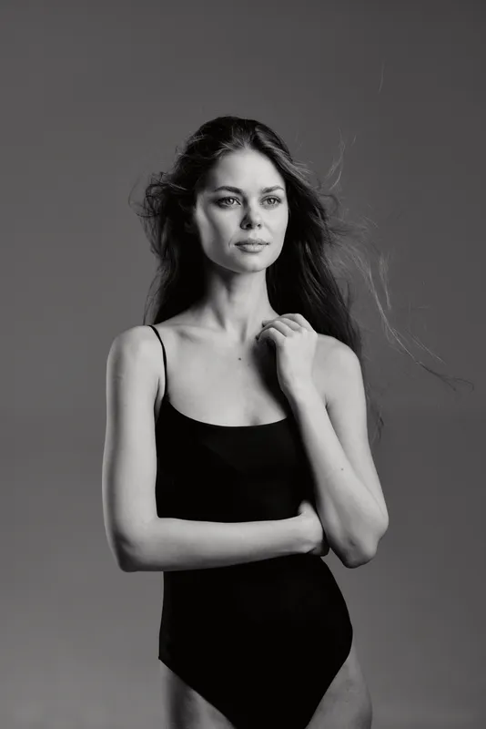Black and white portrait of a contemplative woman in a black sleeveless dress with loose hair on a plain background.