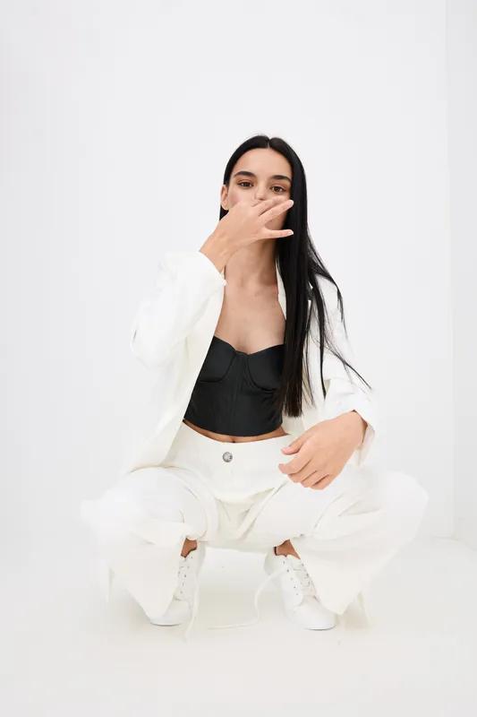 Young woman in white suit and black top squatting with hand near face, isolated on clean white background, fashion pose expressing modern style and confidence.