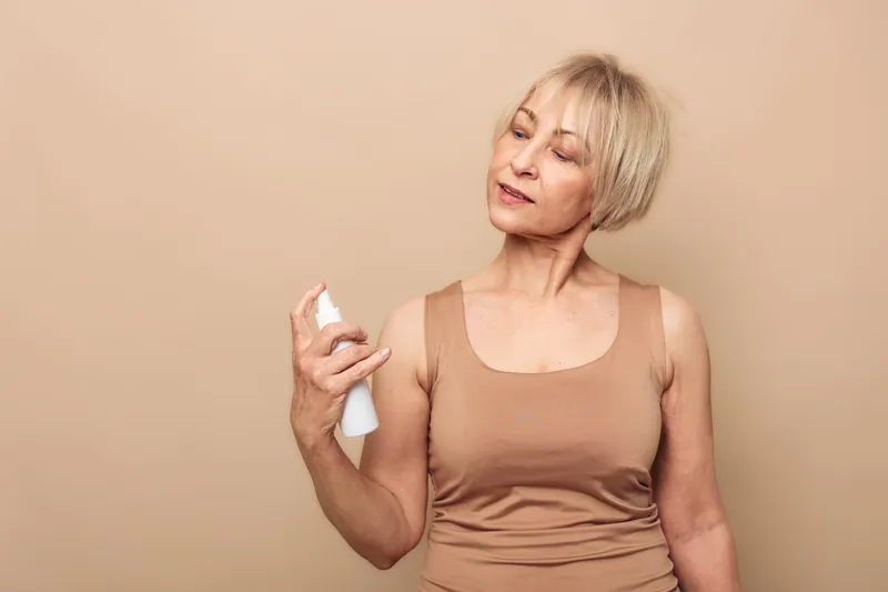 Mature woman with short blonde hair wearing beige top sprays facial mist on her face against a neutral background. Concept of skincare, hydration, and natural beauty for aging skin.