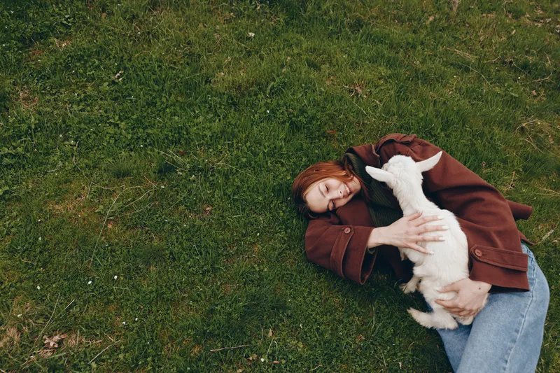 Young woman with red hair lies on green grass holding a white baby goat and smiling happily in a relaxed outdoor lifestyle scene.