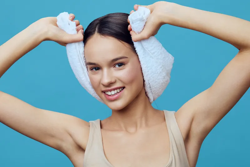Smiling young woman with a towel on her head, showcasing healthy hair and radiant skin against a blue background, radiating freshness and vitality