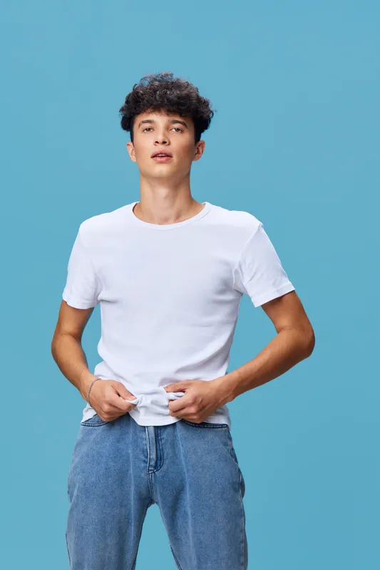 Young man in white t-shirt and blue jeans posing confidently against blue background with casual style and natural expression.