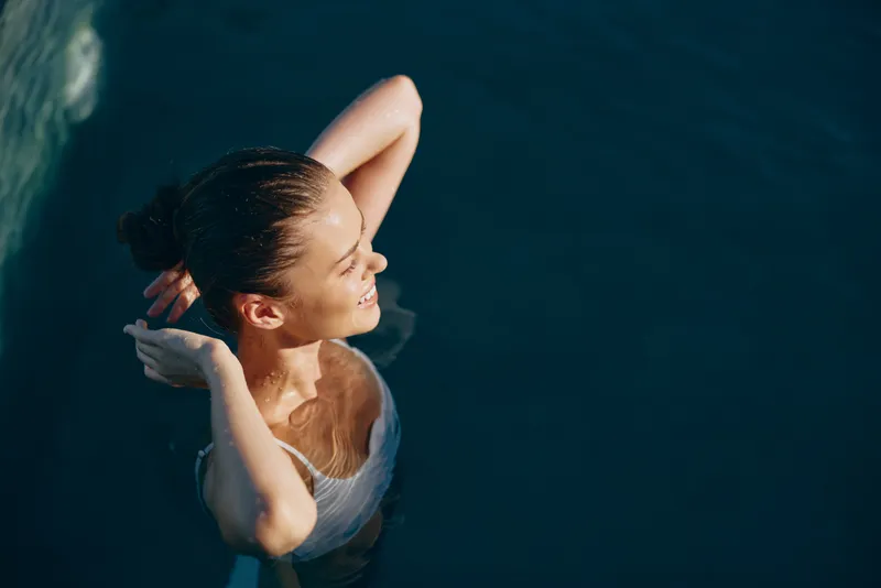 Young woman with wet hair relaxes in a swimming pool with closed eyes and a peaceful smile, enjoying a calm moment of wellness and self-care in natural sunlight.