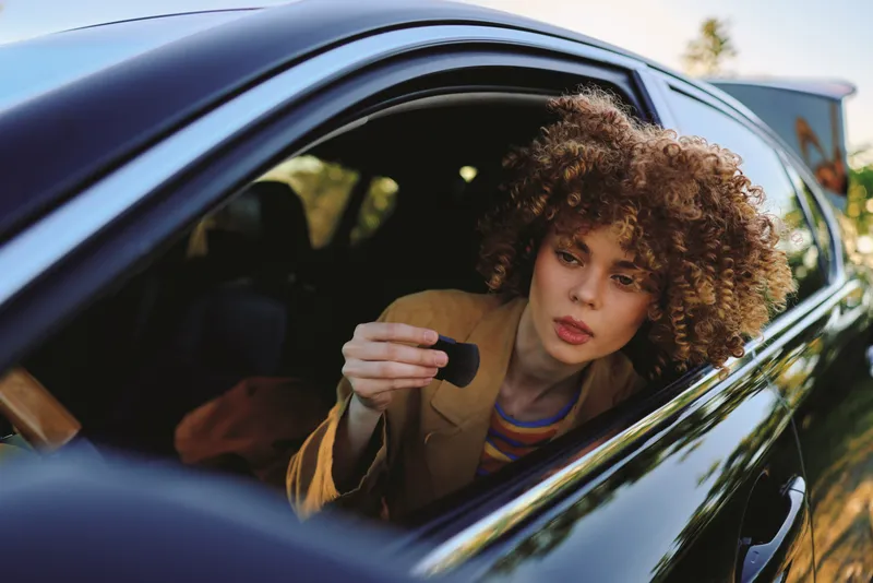 A woman with curly hair in a car applies lipstick, captured in a candid moment through the window. Lifestyle scene with intimate interior, conveying confidence and style.