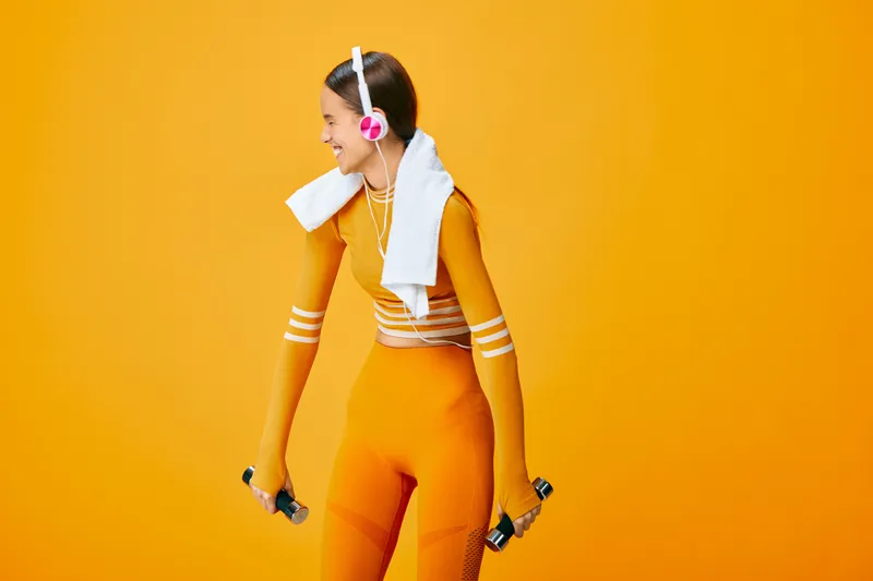 Fit young woman in bright orange activewear holding dumbbells, towel over shoulder and pink headphones, smiling during home workout against solid orange studio background.