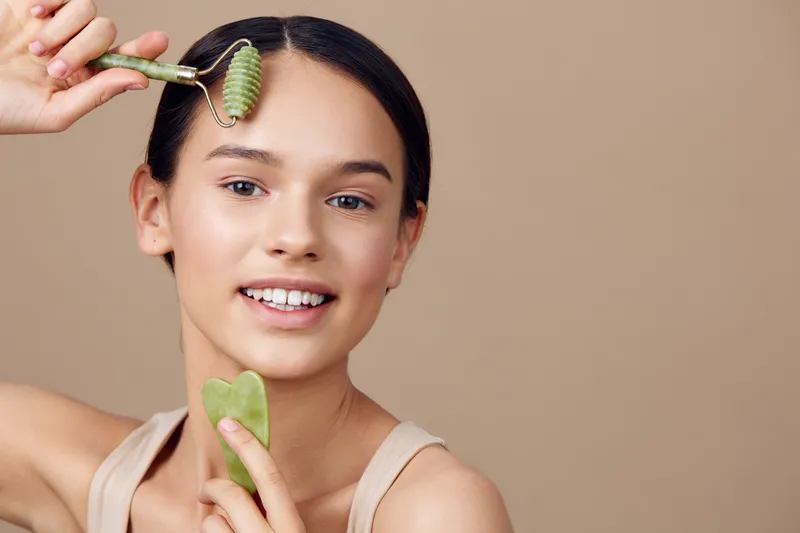Young model using a jade roller on her face, showcasing skincare routine and natural beauty, against a neutral beige background
