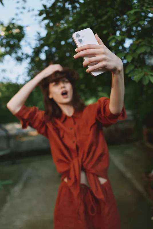Woman in a red dress takes a spontaneous selfie outdoors, capturing a joyful candid moment with a smartphone amid a leafy park setting