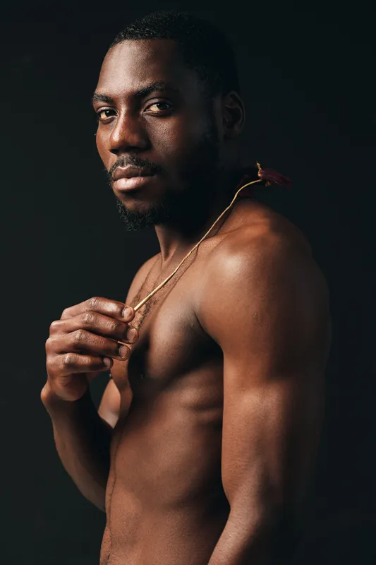 African man with dark skin and muscular body posing shirtless in creative conceptual visual style. Portrait with gold chain, confident expression and studio lighting on black background.