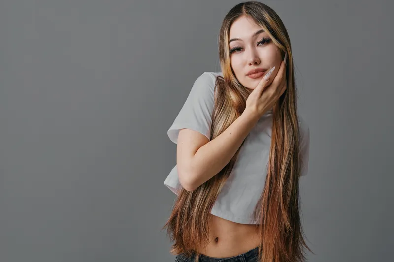 Young woman with long hair posing with hand on face wearing casual crop top against gray background. Beauty, fashion and portrait in studio with natural look and relaxed expression.
