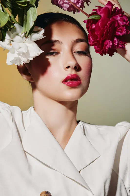 Beauty portrait of a young woman with red lipstick posing with white and red flowers around her face against a muted pastel background. Concept of fashion and creative beauty.