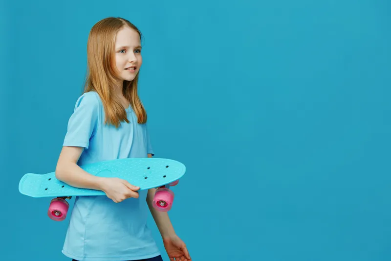Young girl with long hair holding a blue skateboard against a vibrant blue background, showcasing a joyful expression and a casual style perfect for summer fun