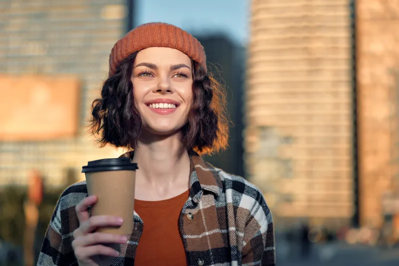 Woman smile coffee urban golden beanie enjoying takeaway cup in sunlit street, candid lifestyle moment with authenticity, golden hour glow, mindful living and emotional storytelling.