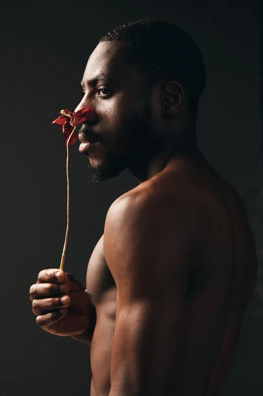 African man with dark skin holding dried flower close to face in creative conceptual visual style. Artistic portrait with bare torso and thoughtful expression on dark background.