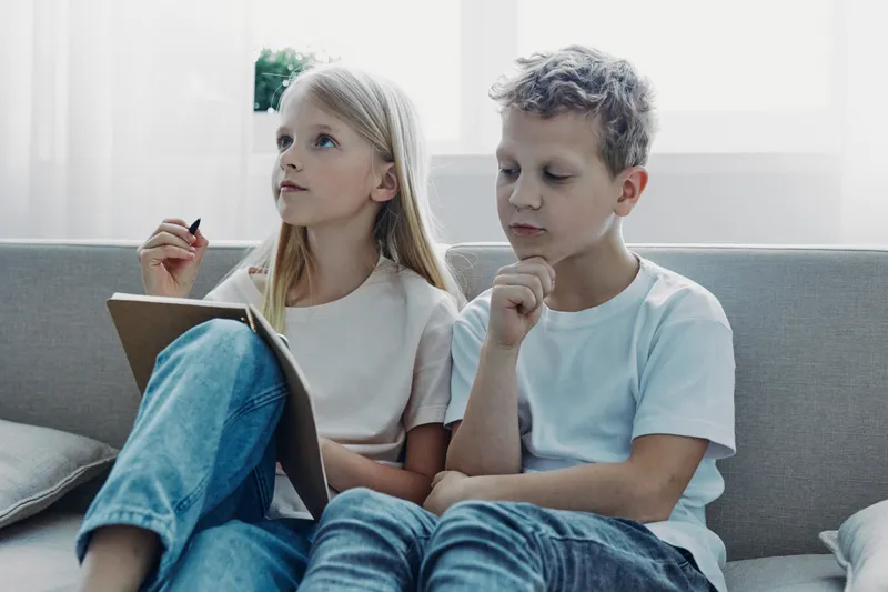 Two children sitting on couch, thoughtful boy and girl with clipboard, indoor light. Concept for learning, concentration, education, and childhood development.