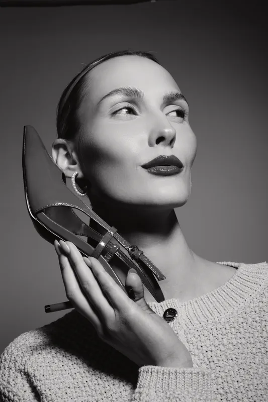 Young white woman with fair skin holds high heel shoe near face wearing knitted sweater in black and white studio portrait.