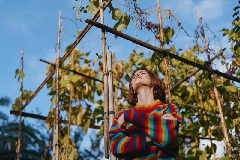 Woman in colorful sweater standing in garden with plants by wooden trellis looking up with folded arms, autumn vines and warm sunlight, contemplative outdoor portrait of seasonal fashion.