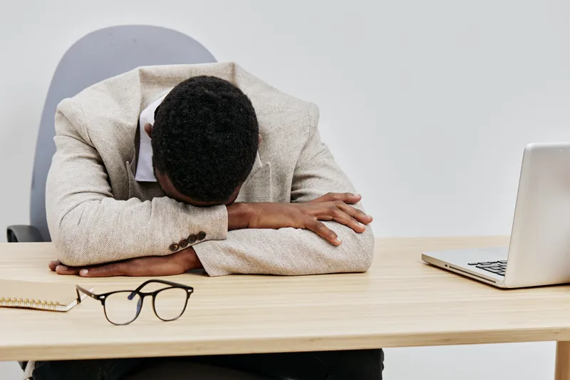 Exhausted young man with curly hair wearing beige sweater and white shirt, resting head on forearms on desk, surrounded by laptop, glasses, notebook, and pen, in modern office interior. People work st
