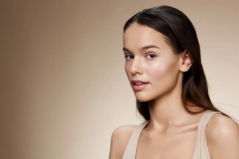 Young woman with long hair in neutral attire, showing a serene expression against a soft beige background, reflecting beauty and simplicity in portrait photography