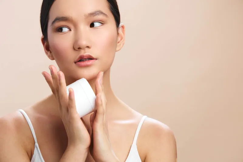 asian woman applies skincare product in a clean studio, serene expression, soft lighting, minimalist setup, showcasing self care, calm confidence, and natural beauty with a tranquil mood.
