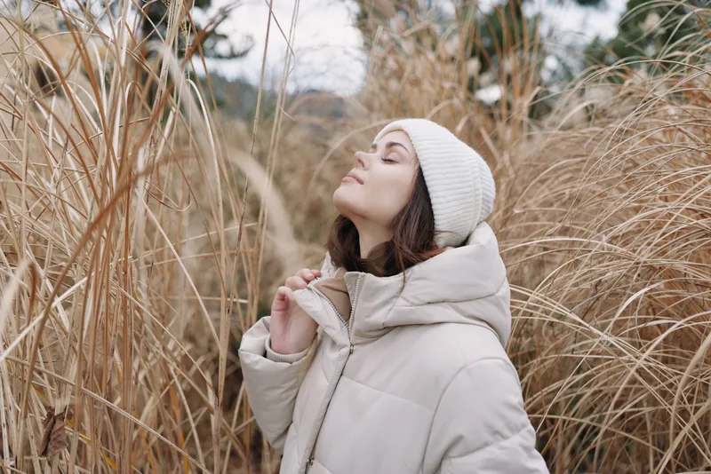 Woman in a warm winter jacket and beanie stands among tall grasses, a calm portrait in natural light, conveying peaceful outdoor mood and cozy seasonal style