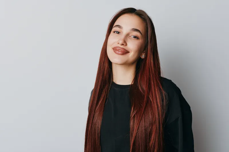 Young woman with long red hair and casual black shirt smiling confidently on light gray background. Portrait of happy female with natural makeup and relaxed expression indoors.