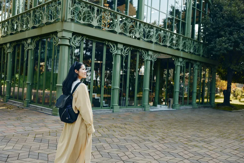 Woman in a long beige coat with a backpack stands beside an ornate glass building in an outdoor urban setting, daylight highlighting fashion and architecture in a thoughtful street scene