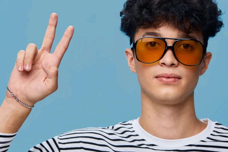 young man wearing orange sunglasses and striped shirt shows peace sign with fingers against blue background, casual style and relaxed expression