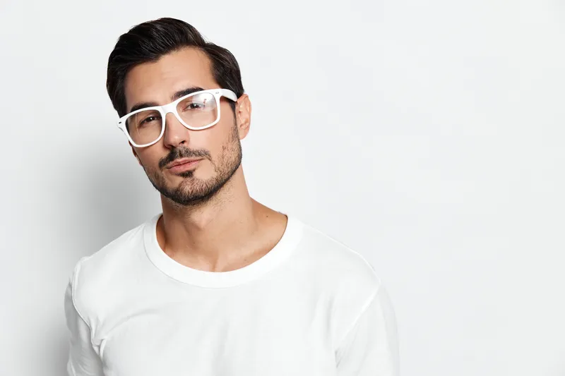 Stylish confident young man wears casual white t-shirt and stylish white glasses, looking at the camera with a serious expression against a plain background.