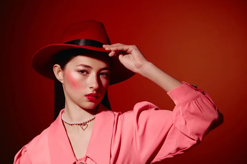 Fashion woman wearing a red hat touches its brim making playful eye contact. She wears pink top with pearls on vibrant red background. Concept of beauty, style, creativity, and elegance.