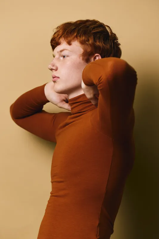 Young redhead man in brown turtleneck sweater posing with hands behind neck against beige background. Side profile with thoughtful expression and casual fashion look indoors.