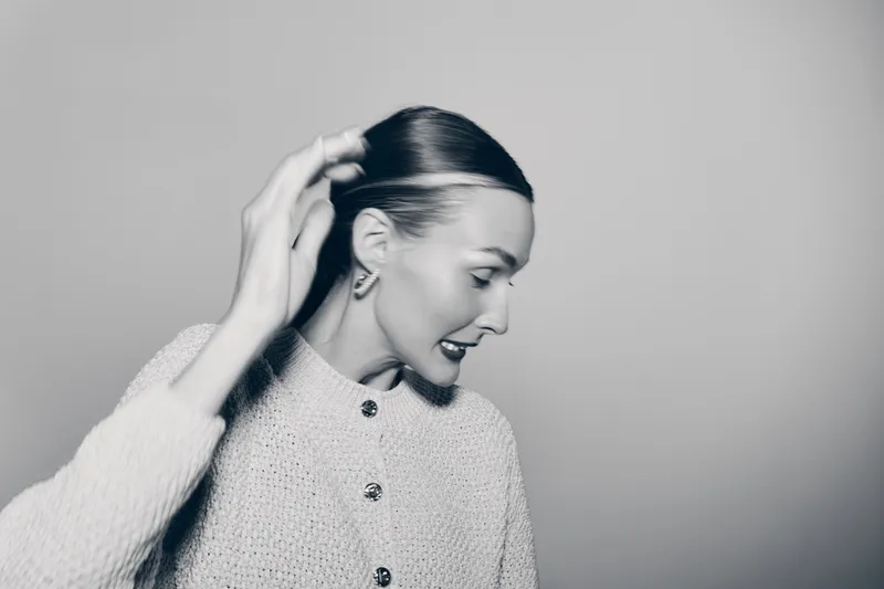 Portrait of a fair skinned caucasian woman in a textured sweater adjusting her hair. Studio lighting highlights her profile against a plain gray background. Concept of fashion and elegance.