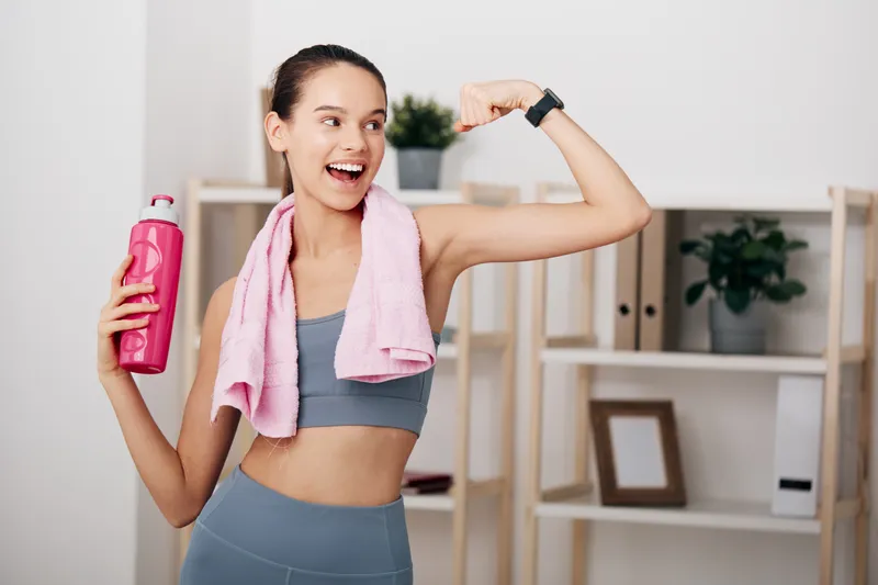 Female athlete in athletic wear flexing her arm after a workout, holding a pink hydration bottle with a towel draped over her shoulders, in a bright home gym with shelves and plants.