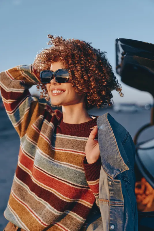 Woman lifestyle curly hair sunglasses denim smile outdoors car, young woman in striped sweater and denim jacket smiles, wearing sunglasses beside parked car, warm autumn sunlight, relaxed cheerful
