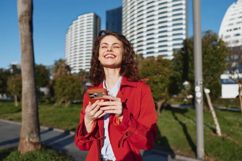 A smiling woman in a red jacket holds a phone in bright sunlight, standing in a city park with modern high rises behind. A lively urban lifestyle moment.