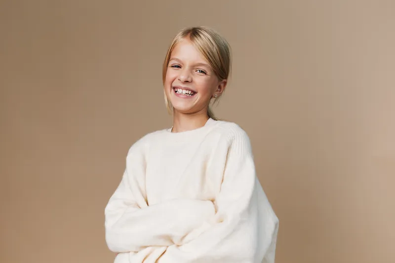 Portrait of a joyful young girl wearing a soft cream sweater, smiling gently with arms crossed. Studio lighting highlights warm tones and a calm, friendly mood.