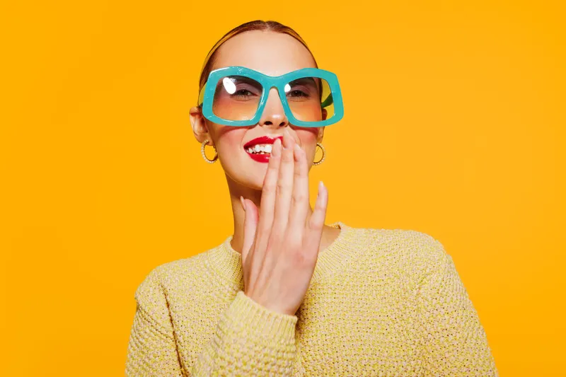Young woman with large turquoise sunglasses and red lipstick laughs covering mouth with hand. Bright yellow background and casual sweater create cheerful, playful mood and fashion concept.