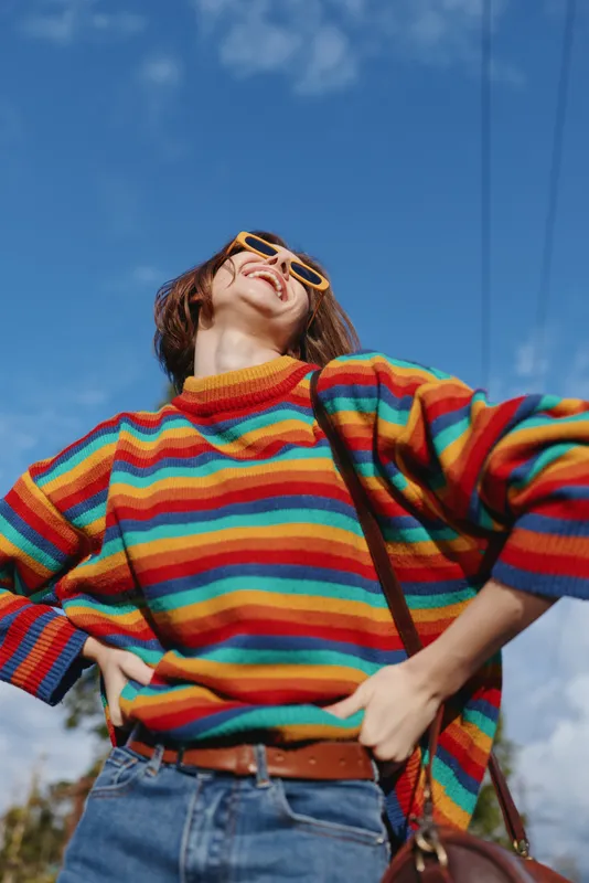 Woman in a colorful striped sweater smiling with yellow sunglasses under a bright blue sky, hands on hips, casual outfit and joyful urban lifestyle portrait full of confidence.