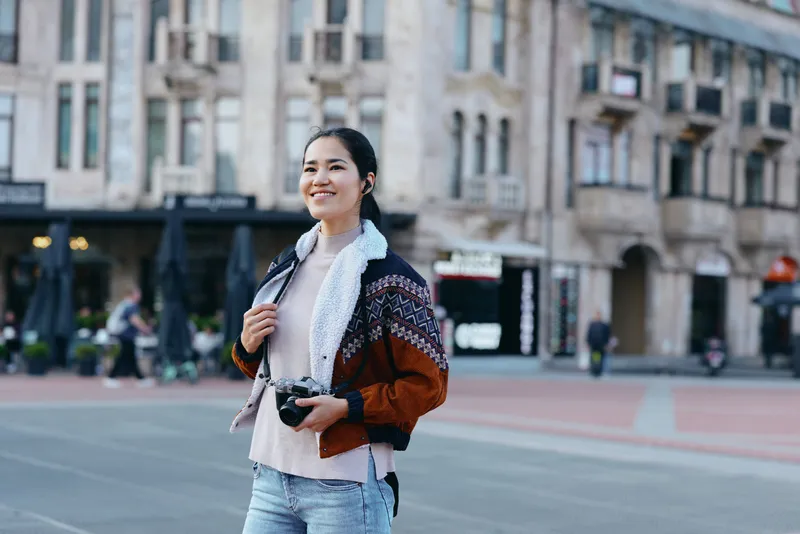 Woman with camera on city street, casual jacket and scarf, urban backdrop, candid street photography showing travel style and everyday exploration