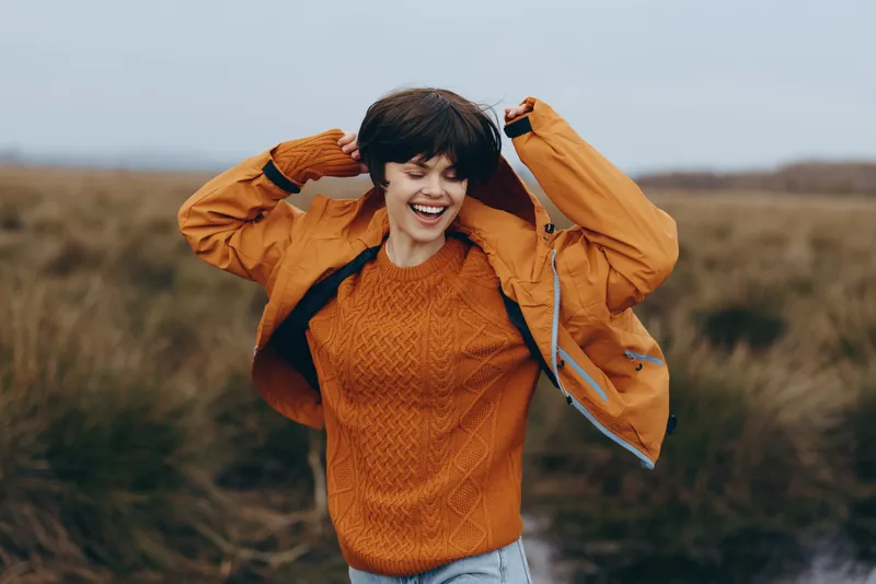 Happy woman lifestyle outdoors in autumn field, smiling, hands on head, wearing orange sweater and jacket, casual clothing, warm nature background, countryside landscape, friendly portrait