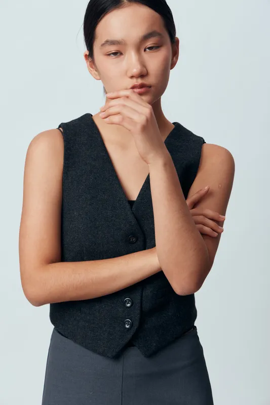Young woman with natural look wearing black vest and dark pants posing with arms crossed and hand near face against plain light background in studio portrait.