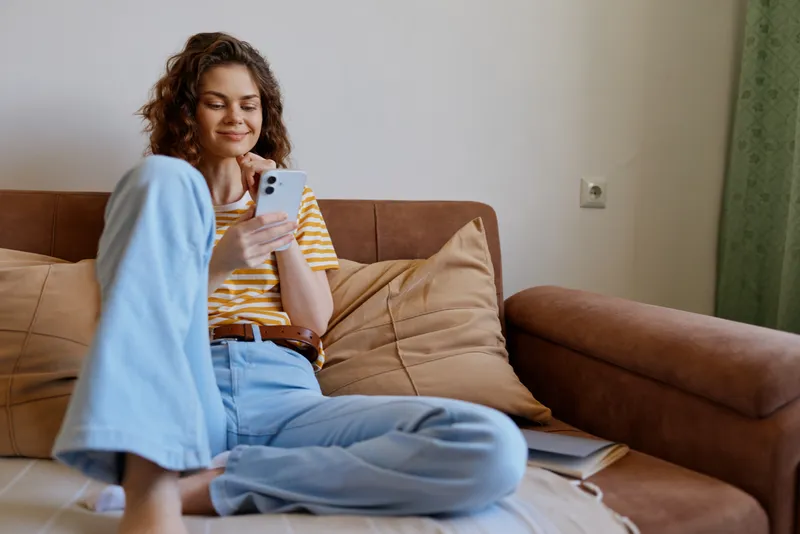 A woman relaxing at home on a couch, checking her phone, embodying casual lifestyle, comfort, and livingroom calm in a bright interior setting, intimate everyday moment