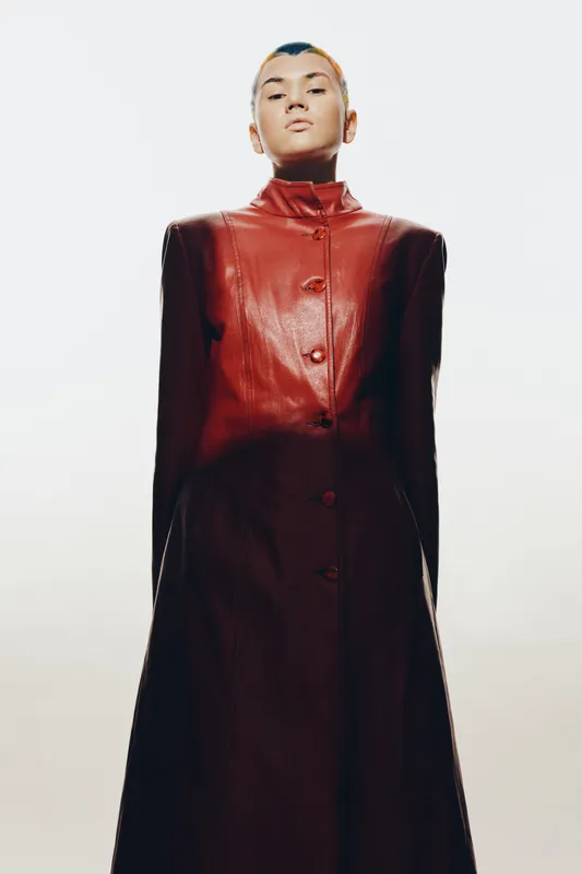Woman wearing a dark red leather coat standing confidently against a white background with minimal shadows and a strong upward camera angle.
