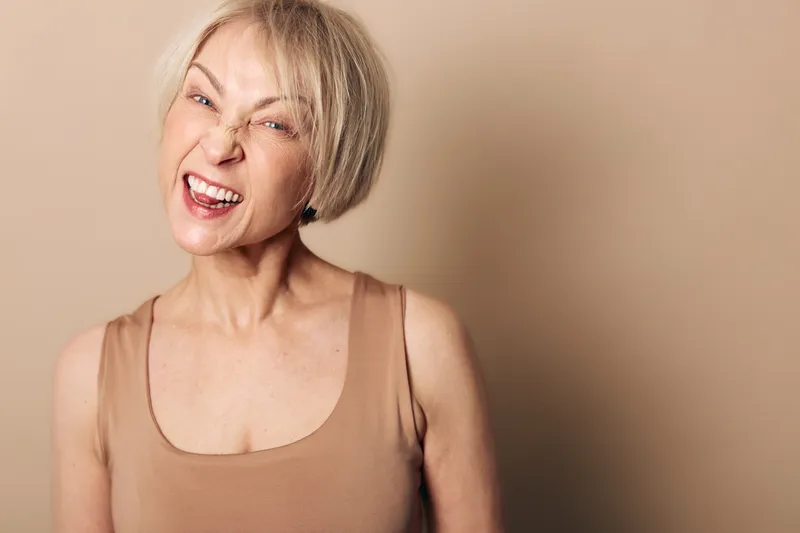 Blonde woman with short hair making a funny grimace stands against beige background wearing sleeveless top. Concept of humor, personality, expression, and casual lifestyle portrait.