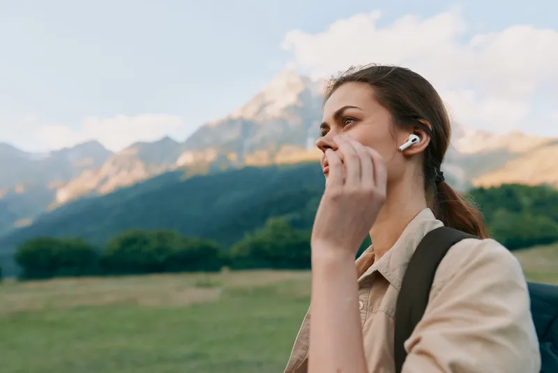 An outdoor portrait shows a young woman with a beige shirt and backpack listening with wireless earbuds against a backdrop of mountains.
